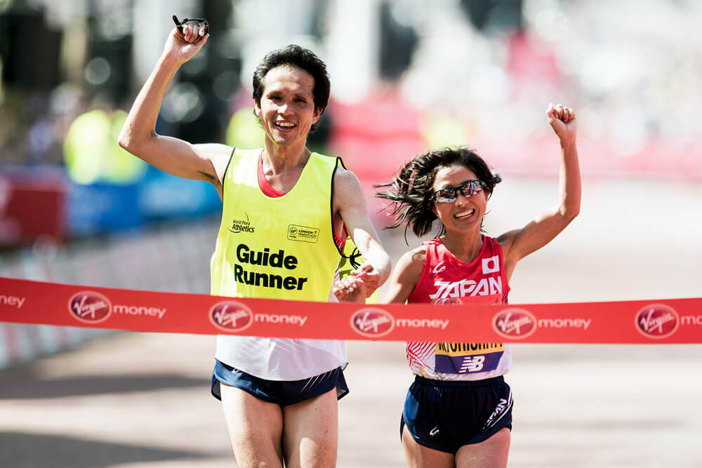 写真：道下美里選手（陸上）が伴走者とともに笑顔でゴールテープを切る瞬間。互いに繋いでいない方の手をガッツポーズするかのように掲げながらのゴール。