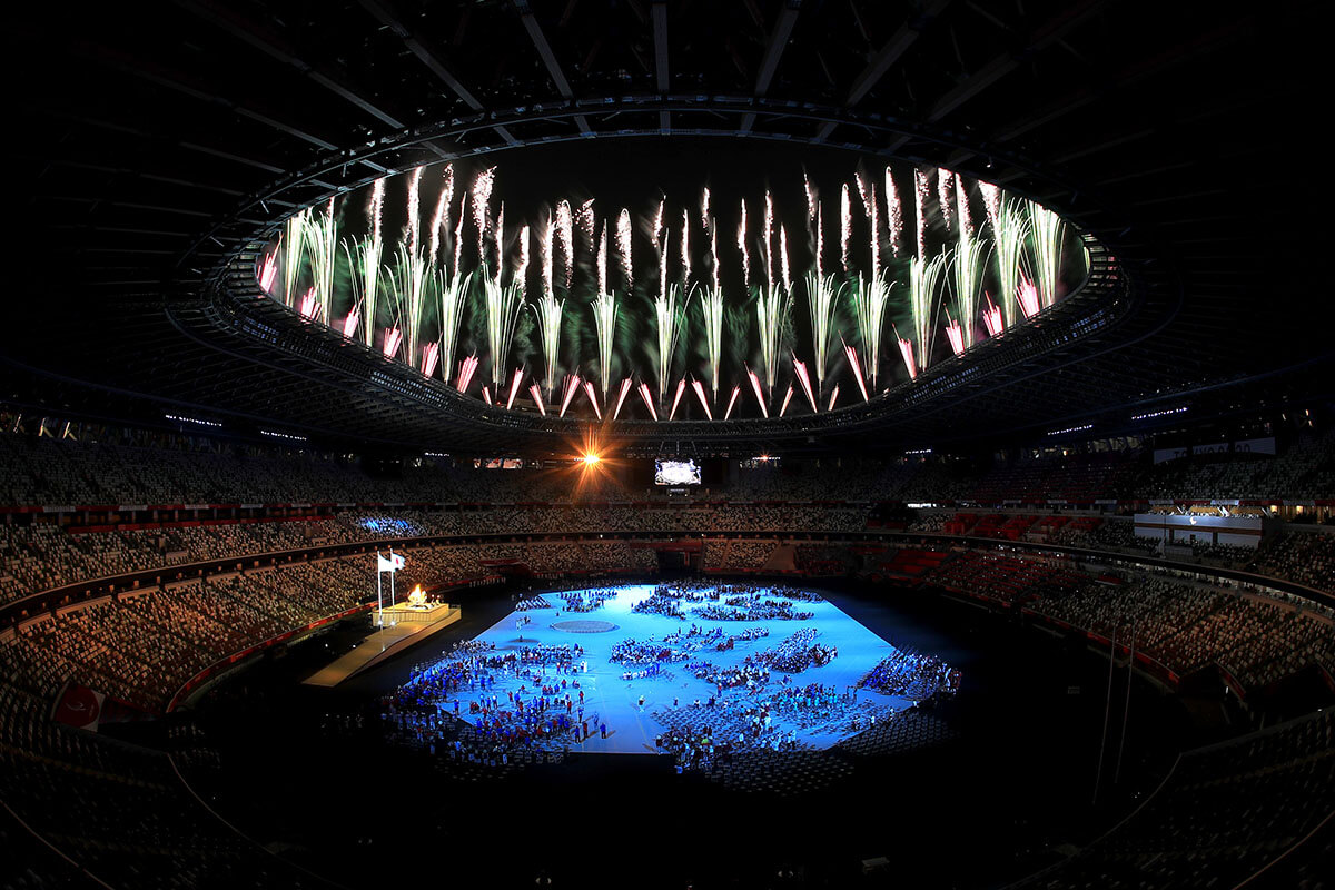 写真：パラリンピックの聖火が灯された直後の開会式会場。選手たちがいる競技場は青く照らされ、その真上に花火が上がっている。
