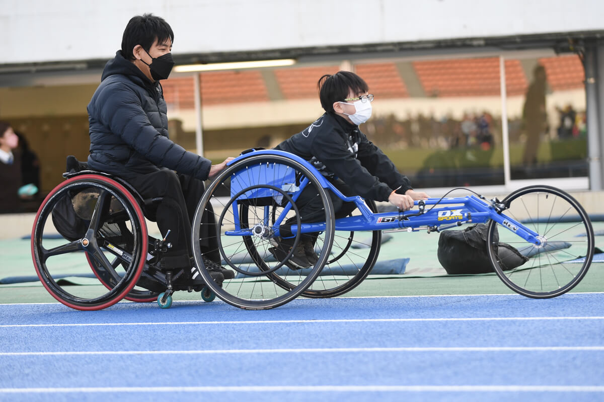 写真：競技用車いすに乗る子どもと、それを支え見守る車いすの大人